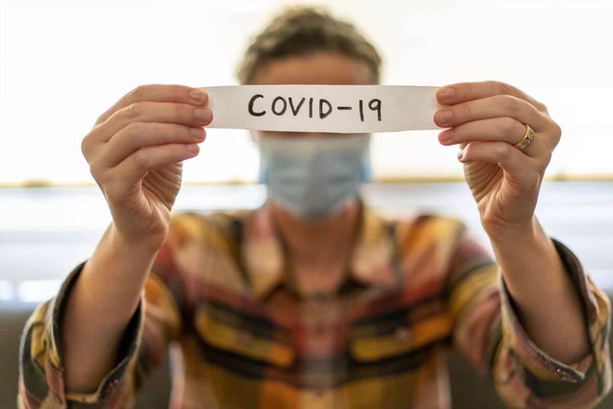 Person wearing mask holding COVID-19 sign, indoor healthcare setting, pandemic awareness.