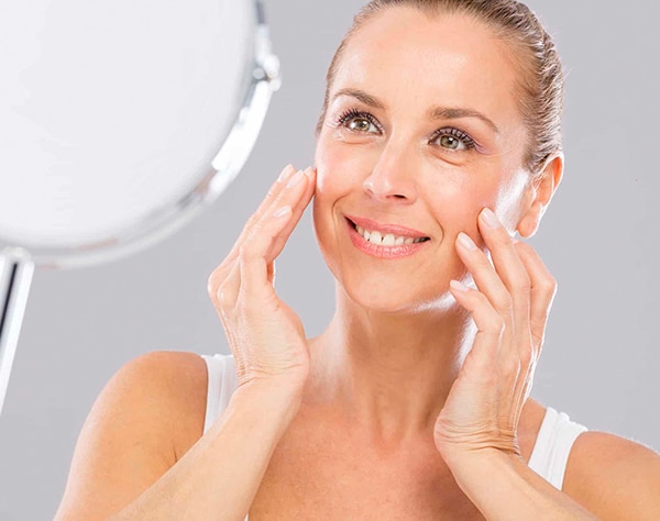 Woman examining her skin in mirror, touching cheeks, bright clinical bathroom setting.
