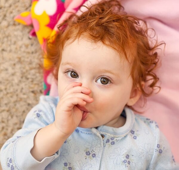 Toddler with curly red hair sucking thumb while lying on pink blanket indoors.
