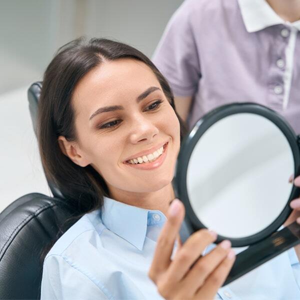 Woman in dental chair holding mirror, smiling, with dentist in background at Brighton Healthy Smiles.