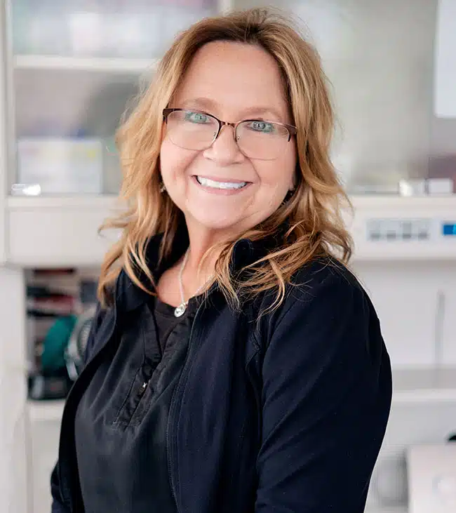 Woman with glasses smiling at camera in bright dental office setting.