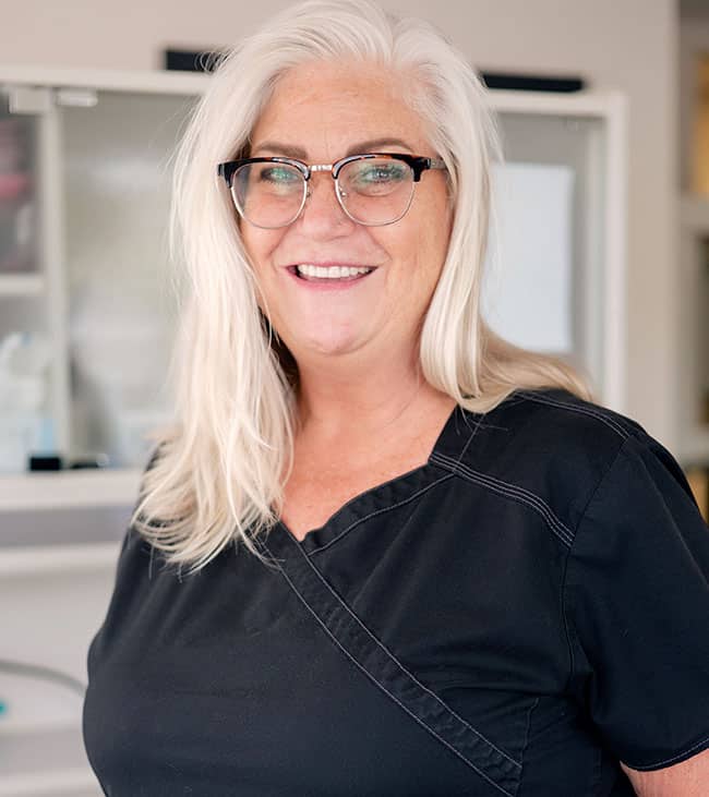 Woman with white hair and glasses smiling in dental office setting.