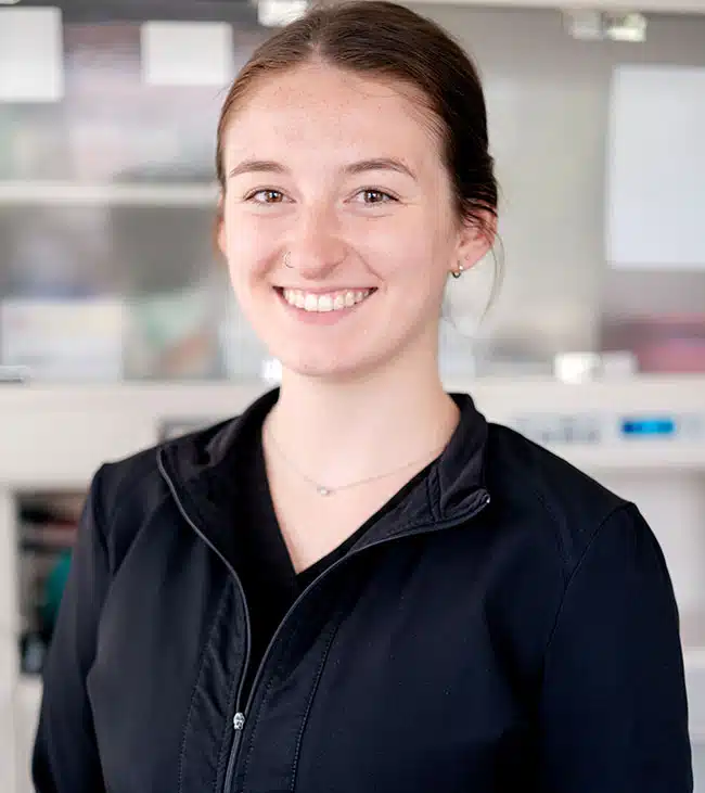 Young woman smiling at camera in professional dental office setting, wearing dark jacket.