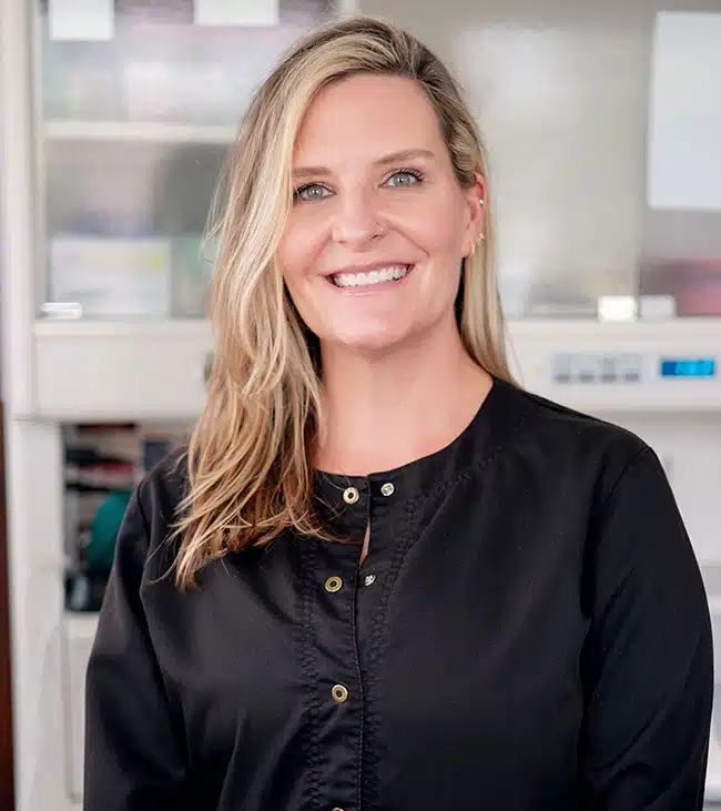 Professional headshot of smiling woman in black blouse against blurred office background.