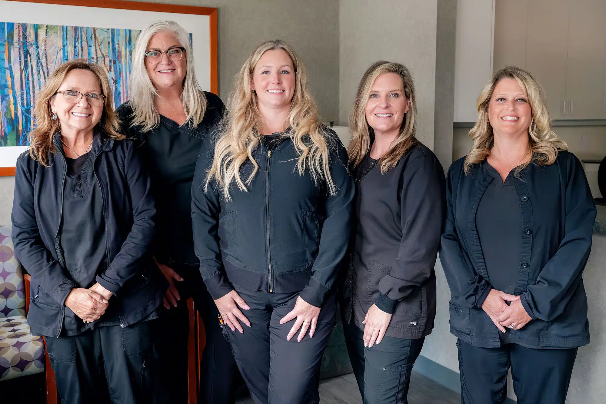 Five female dental professionals in dark uniforms posing together in a modern office setting.