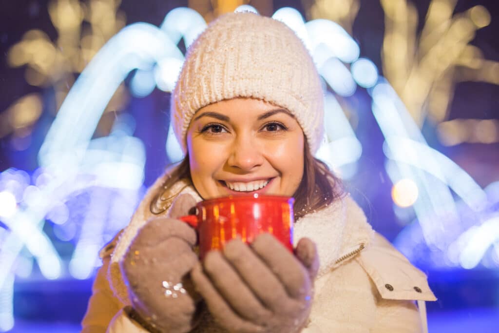 A woman in a white knit hat and coat smiles warmly, holding a red mug. Colorful holiday lights blur in the background, creating a festive atmosphere.