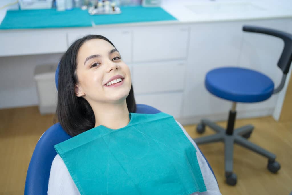 A woman smiling in a dental chair, wearing a teal bib. The room is clean and bright, with dental equipment in the background, creating a calm atmosphere.