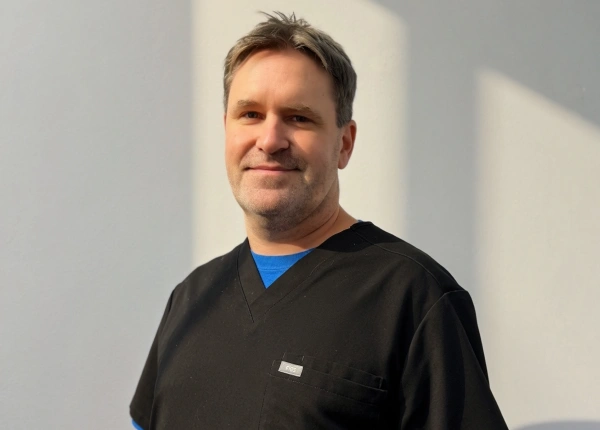 Close-up portrait of Dr. John Hoitenga smiling gently, wearing black medical scrubs over a blue undershirt against a plain white background.